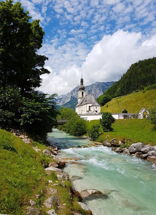 Kirche in Ramsau vor einer Bergkulisse und dem Flüsschen Ramsauer Ache im Vordergrund