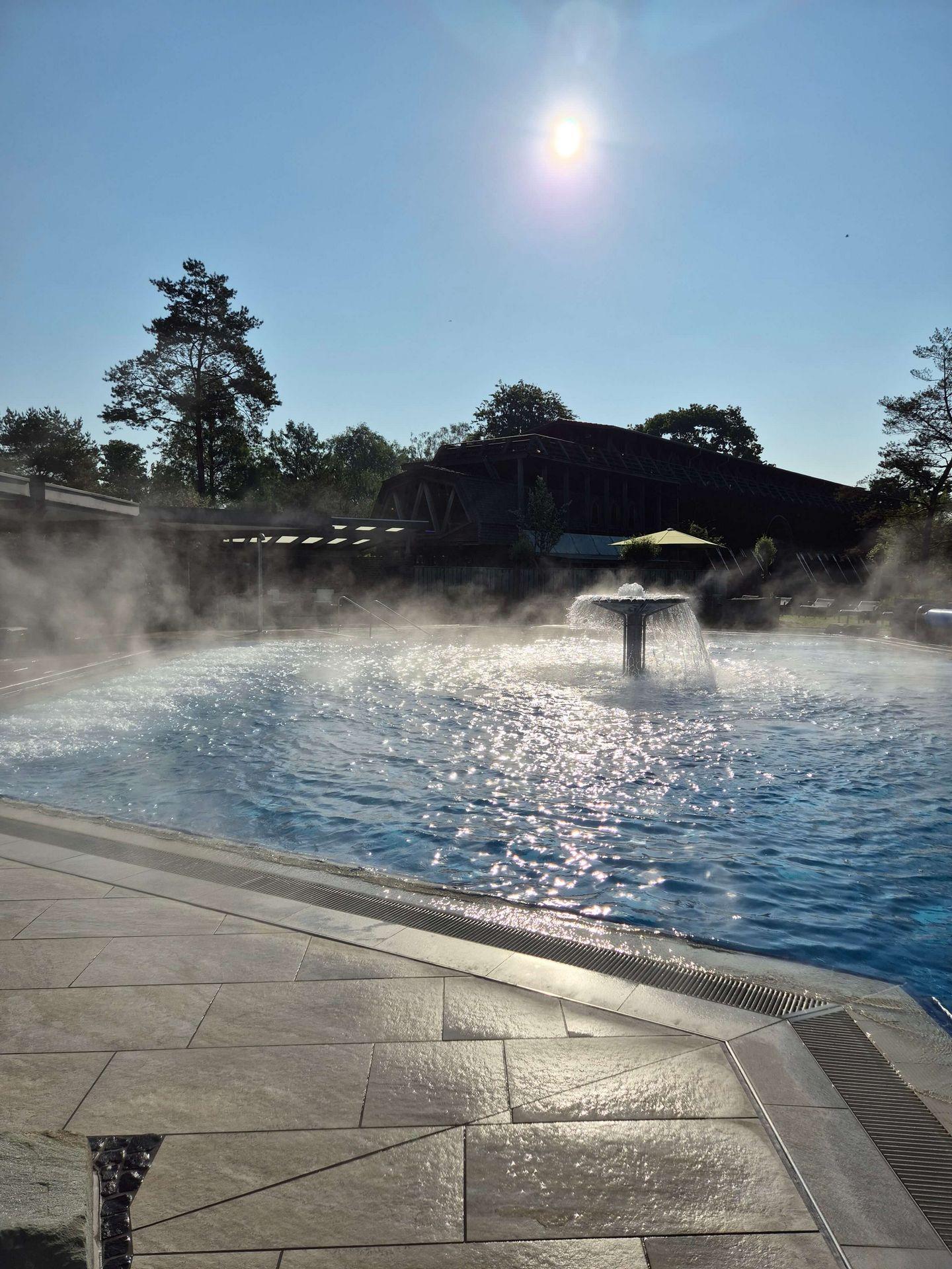 Dampfendes Außenbecken der Börde-Therme in Bad Sassendorf mit Wasserfontäne bei strahlendem Sonnenschein.