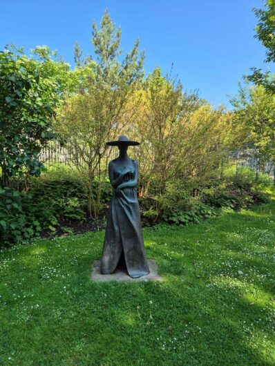 Dunkle Skulptur einer stehenden Frau mit breitem Hut und langem Kleid auf einer grünen Wiese im Kurpark Bad Sassendorf.