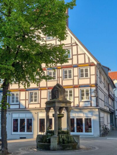 Historisches Fachwerkhaus mit heller Fassade und bunten Holzbalken hinter einem steinernen Brunnen in der Altstadt von Soest.