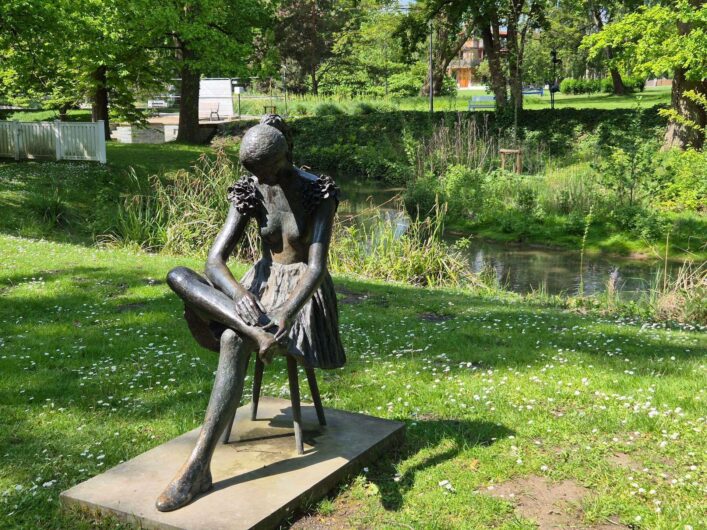 Bronzeskulptur einer sitzenden Frau, die ihren Fuß betrachtet, auf einer Wiese am Wasserlauf im Kurpark Bad Sassendorf.