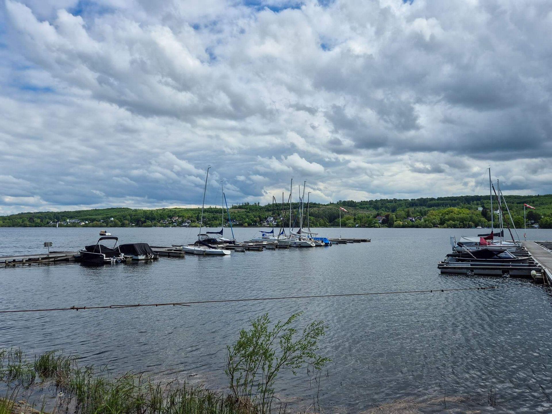 Segelboote und Motorboote an den Stegen eines Yachthafens am Möhnesee mit bewaldeten Ufern und wolkenreichem Himmel.