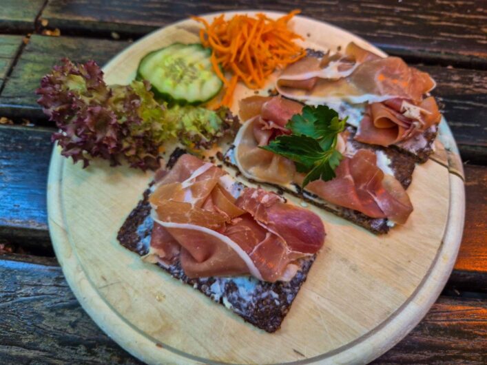 Zwei Scheiben westfälischer Pumpernickel mit luftgetrocknetem Schinken auf einem Holzteller, garniert mit Salat, Gurke und Möhren.