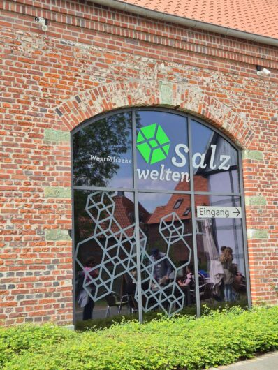Großes Bogenfenster der Westfälischen Salzwelten in Bad Sassendorf mit Museumslogo, Salzkristall-Motiven und Eingangshinweis.