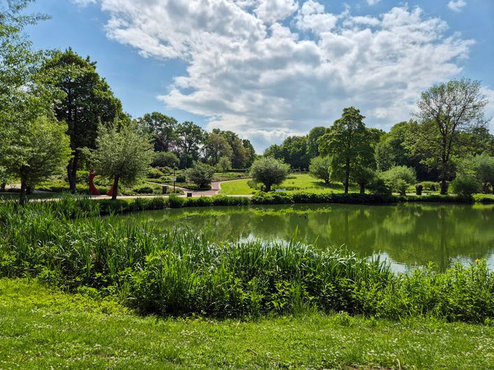 Idyllischer See im Kurpark Bad Sassendorf mit grünen Uferpflanzen, weitläufigen Rasenflächen, Spazierwegen und hohen Bäumen.